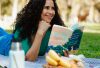 Smiling woman reading a book at a picnic in a sunny park setting.